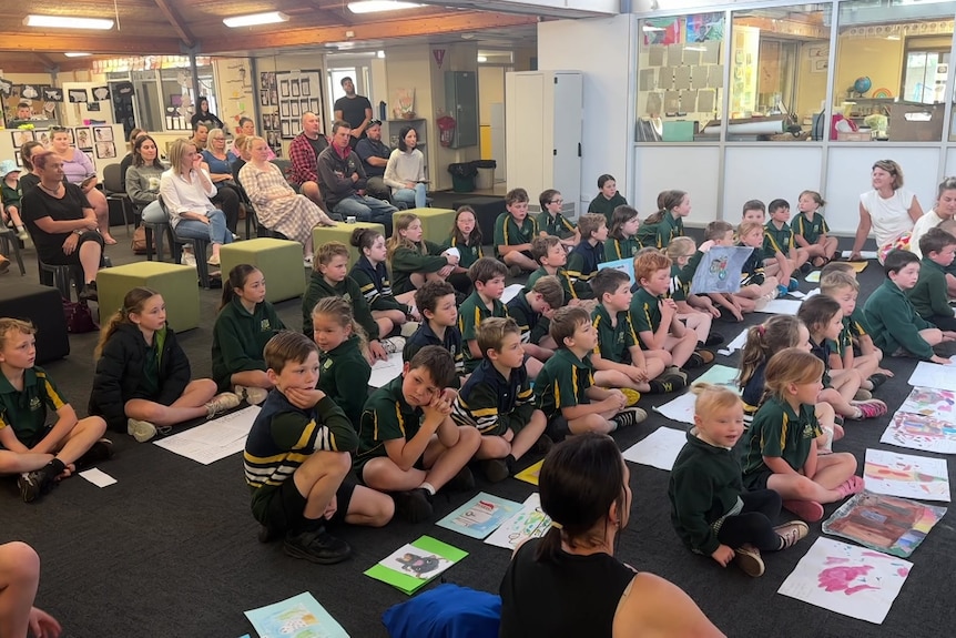 Group of children sitting on the floor with paper in front of them, adults sitting at back watch on.