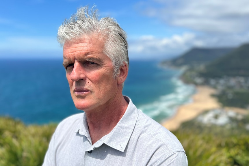 An older man with short grey hair wearing a collared shirt stands on a cliff with a sunny beach behind him.