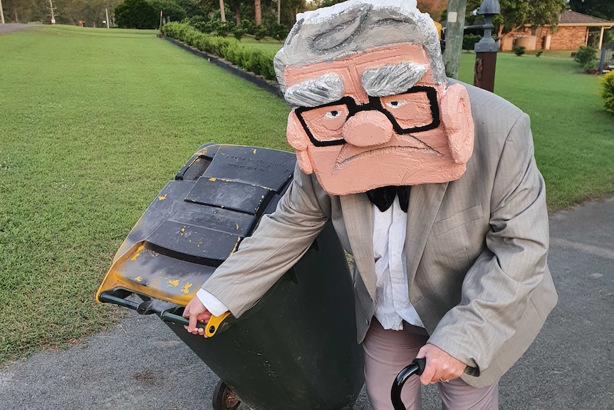 A woman wearing a costume of a large styrofoam head of an old man wheels her bin to the kerb.