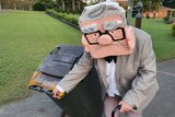 A woman wearing a costume of a large styrofoam head of an old man wheels her bin to the kerb.