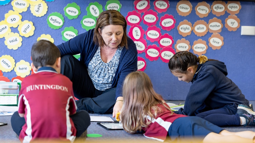 A teacher sits on the flood in a classroom, showing three students writing.