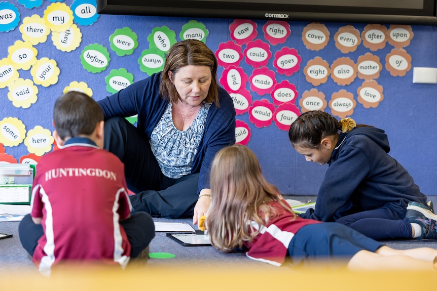 A teacher sits on the flood in a classroom, showing three students writing.