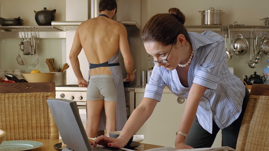 Man wearing only underpants and apron cooking while older woman uses laptop at the table in a kitchen