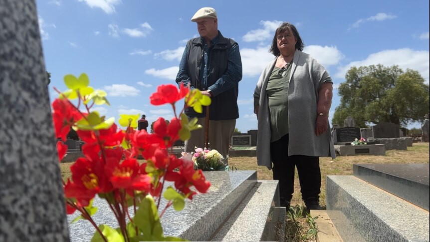 a man with a cane and hat and woman wearing a grey cardigan look at a gravesite. red flowers in the foreground