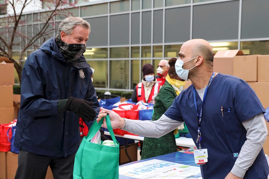 NYC Mayor Bill de Blasio in gloves with scarf on his face hands a bag to a medical worker in face mask and scrubs.