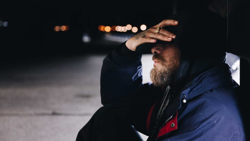 An anonymous  man holds his forehead while sitting in a street at night.