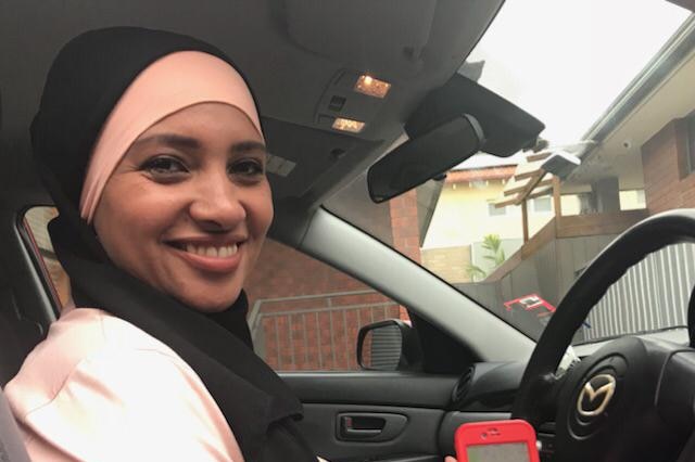 A smiling woman wearing a head covering holds up a phone while she sits behind the wheel of a car.