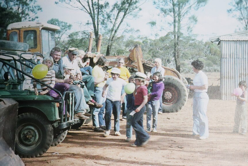 People, children sitting on vehicles, grader including with balloons, tress in background, shed on right 