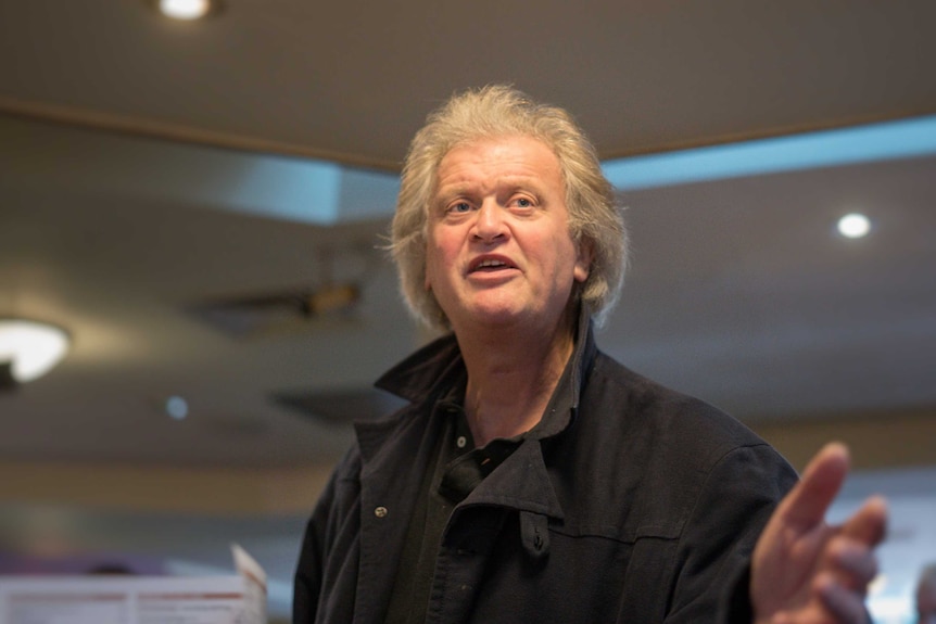 A man with blonde hair wearing a black jacket speaks to a crowd.