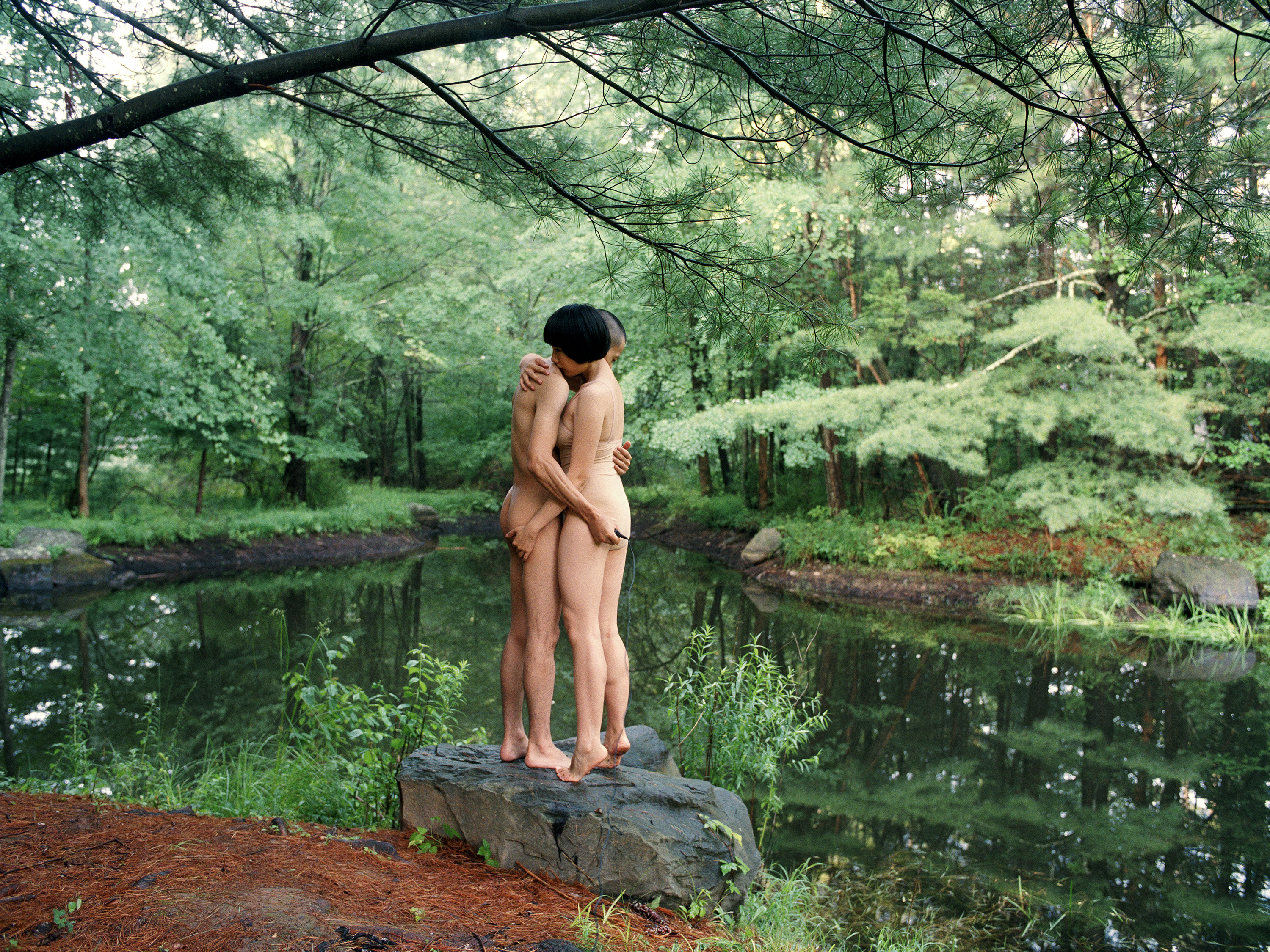 Pixy Liao, a Chinese woman with short black hair, and Moro, a Japanese man with a shaved head, hug naked by a forest green pond