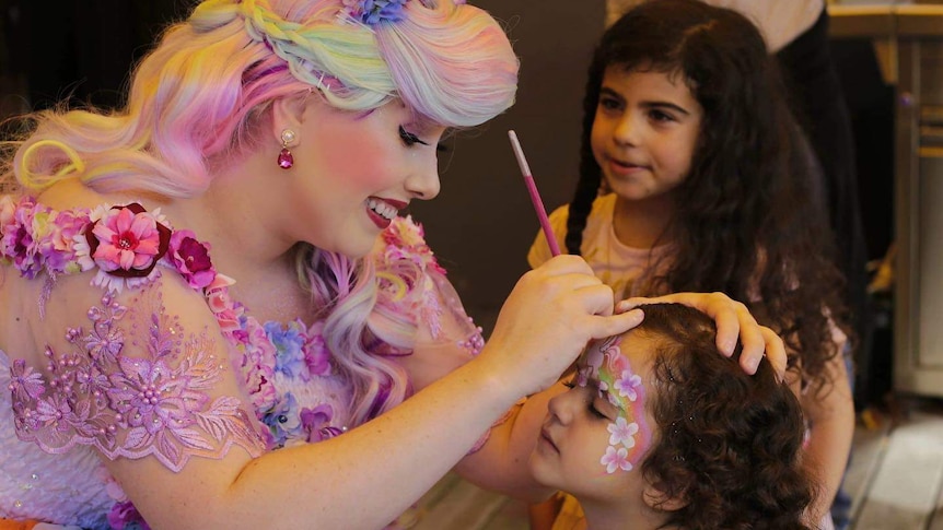 Emily Turnbull is dressed as some kind of unicorn princess and paints a child's face at a party.