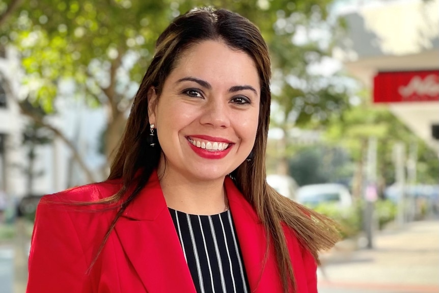 woman in red jacket with brown hair and brown eyes smiling in front of green trees