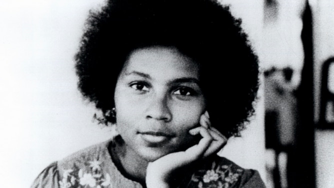 A woman with a dark afro rests her head in her palm as her photo is taken, in black and white