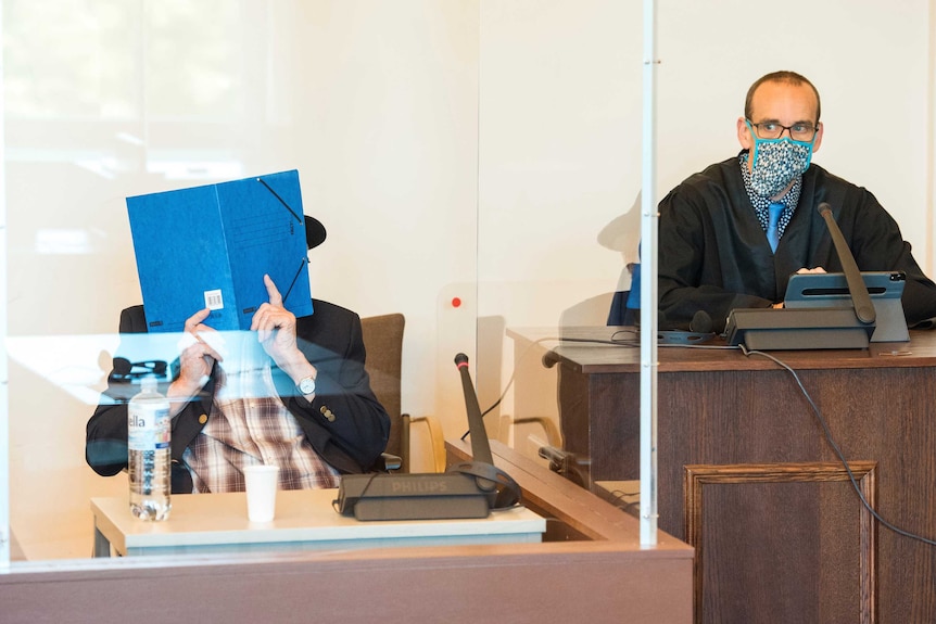 An elderly man sits in a wheelchair on the stand. Separated by perspex, the judge sits next to him wearing a mask.