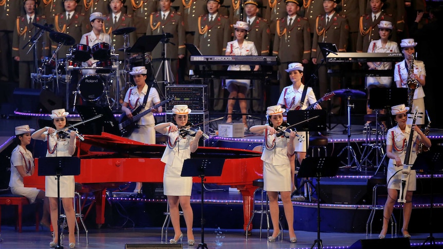 North Korean musicians dressed in white and green suits stand on stage playing their instruments.