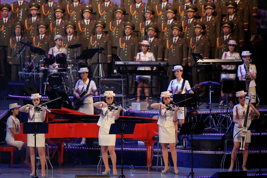 North Korean musicians dressed in white and green suits stand on stage playing their instruments.