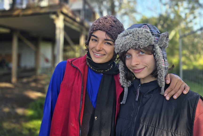 Two smiling young women, dressed for warmth, stand near a house.