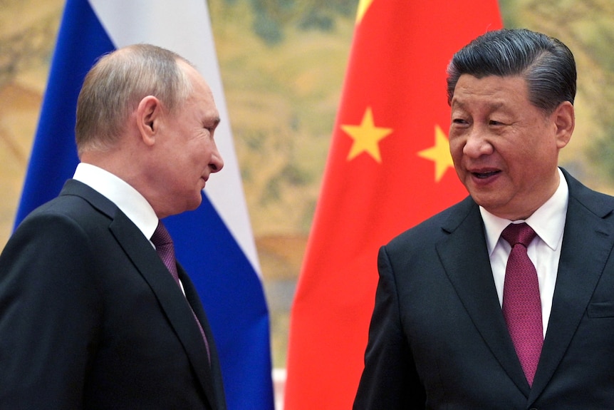 Vladimir Putin smiles as he shakes hands with Xi Jinping in a room lined with flags.