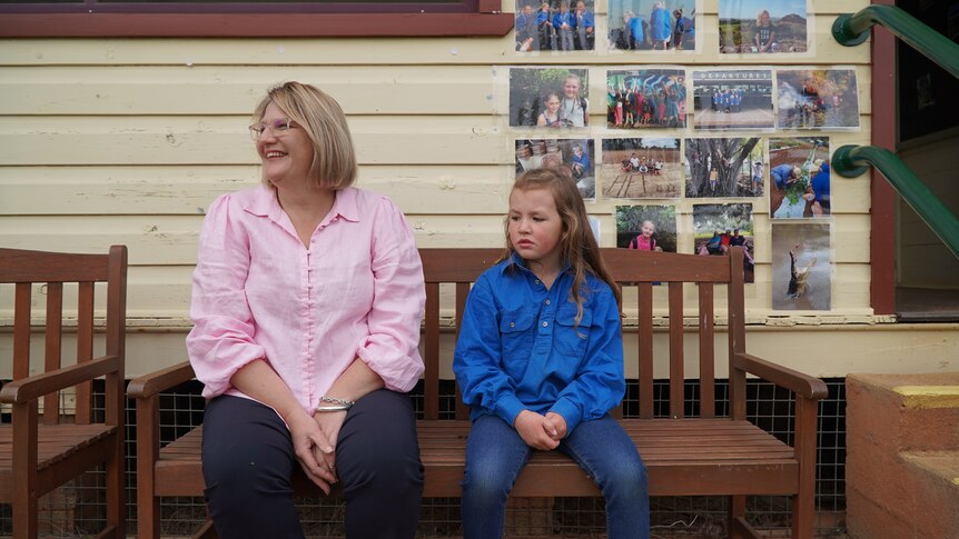 A blonde woman in a pink shirt and a primary school child sit on a park bench outside a schoolhouse.
