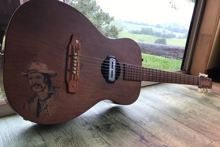 A guitar by a window, with brown wood and a self-portrait on the wood.