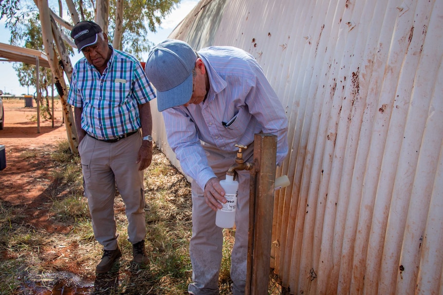 Two men stand at tap pouring water into a bottle in a remote Indigenous community.