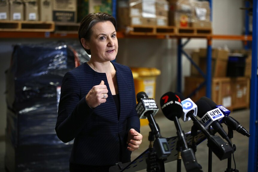 A middle-aged woman with medium length brown hair and a navy blazer holds a press conference in a warehouse.