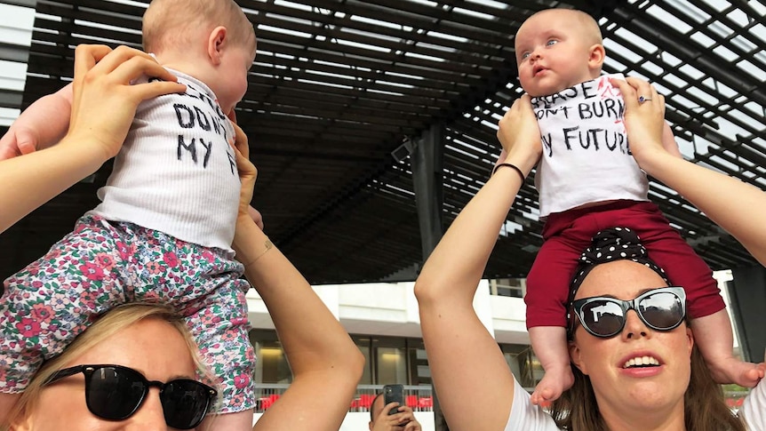 Two mothers hold their babies on their shoulders wearing protest singlets at the Brisbane climate rally.