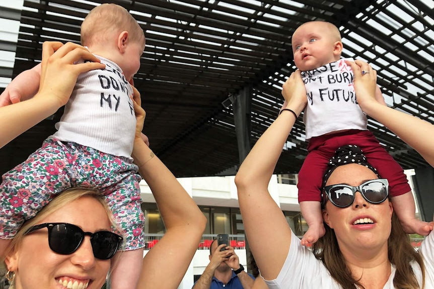 Two mothers hold their babies on their shoulders wearing protest singlets at the Brisbane climate rally.