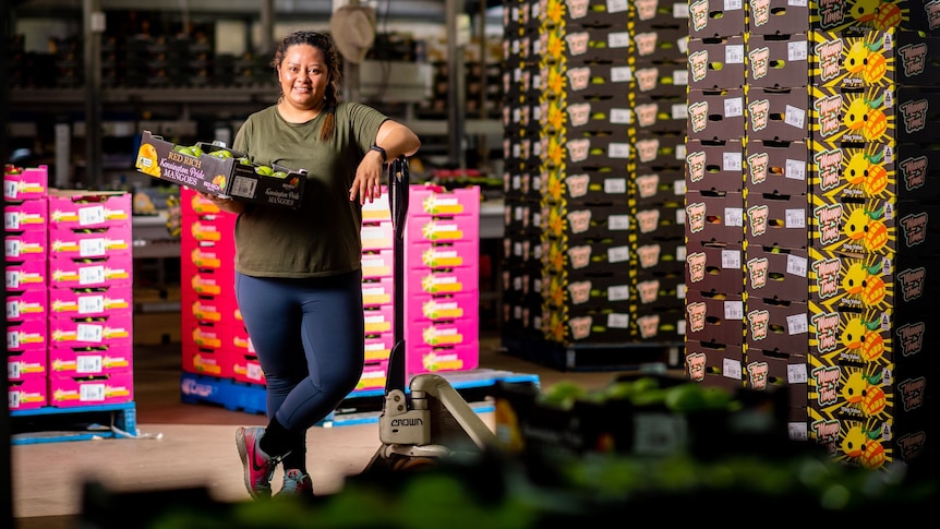 A young Samoan woman smiles in a warehouse carrying a box of mangoes