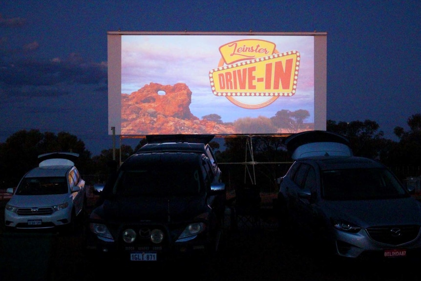 A drive-in movie theatre screen with cars parked in front of it.