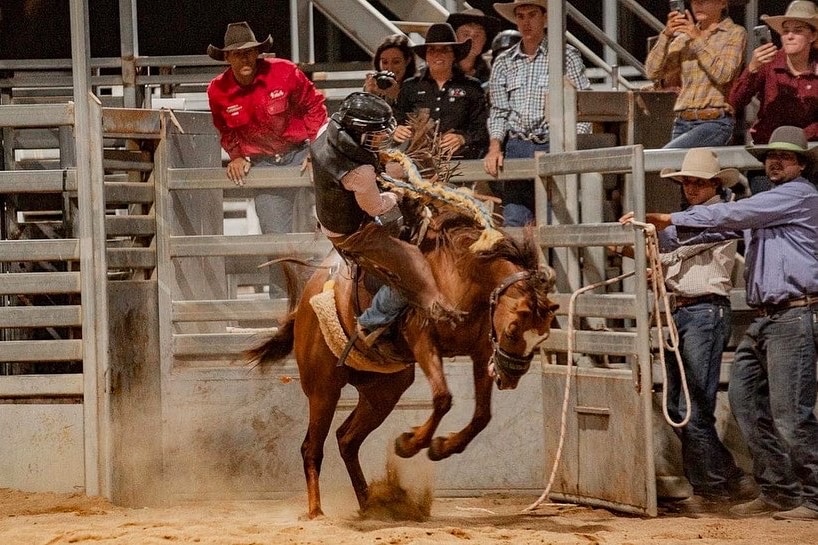 Onlookers lean on a stock fence as a woman begins to fall off a bucking horse.