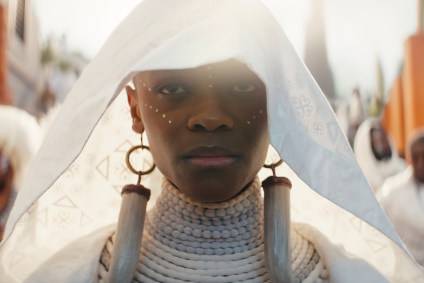 A close-up of a black woman wearing a white veil, large ivory and gold earrings and stacked white beaded necklaces.
