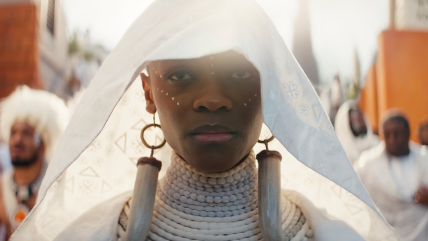 A close-up of a black woman wearing a white veil, large ivory and gold earrings and stacked white beaded necklaces.