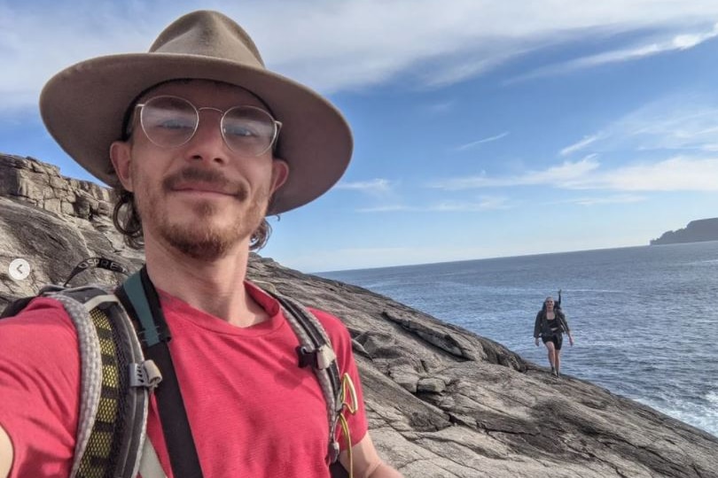 A young man takes a photo of himself at a cliff face, smiling.