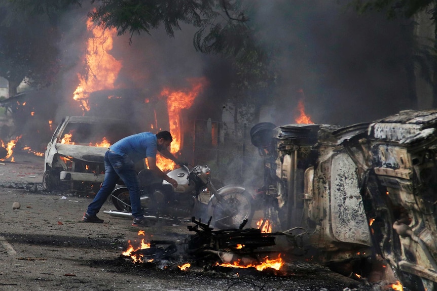 A man lifts a motorbike off the ground as burning cars blaze behind him.