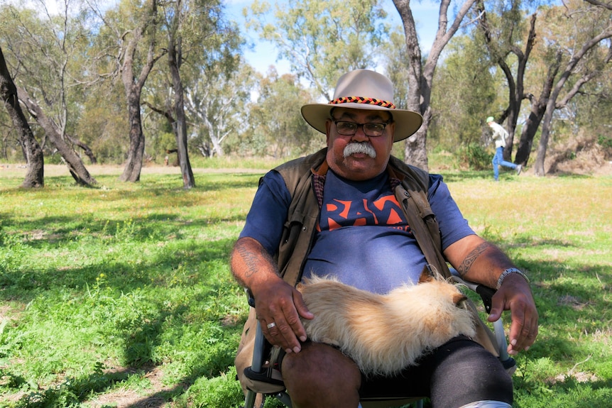 A man sitting under the trees with a small dog on his lap 