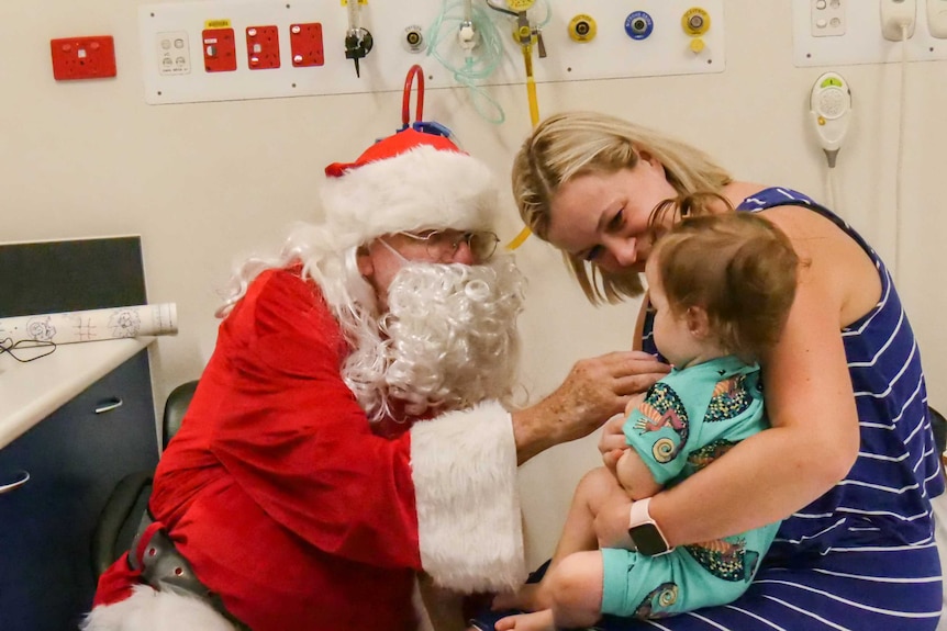 A man dressed as Santa talks to a baby girl, held by her mum.