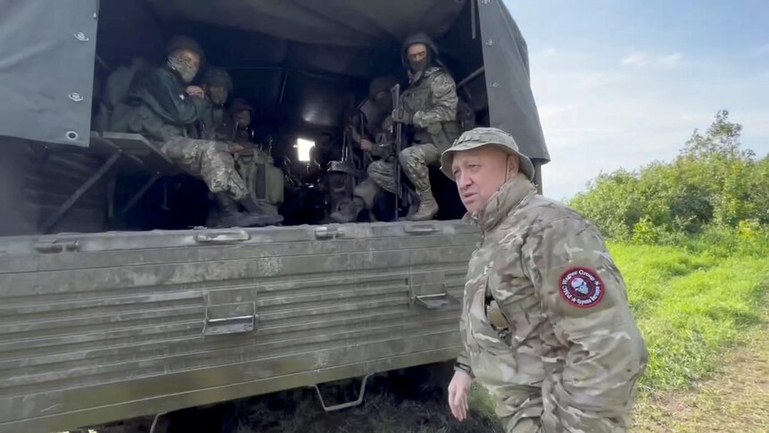 The back of an army truck with soldiers is pictured, in front of it stands Wagner chief Prigozhin.
