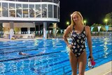 A picture of a smiling swimmer standing next to a pool with lights on and people swimming in the background.