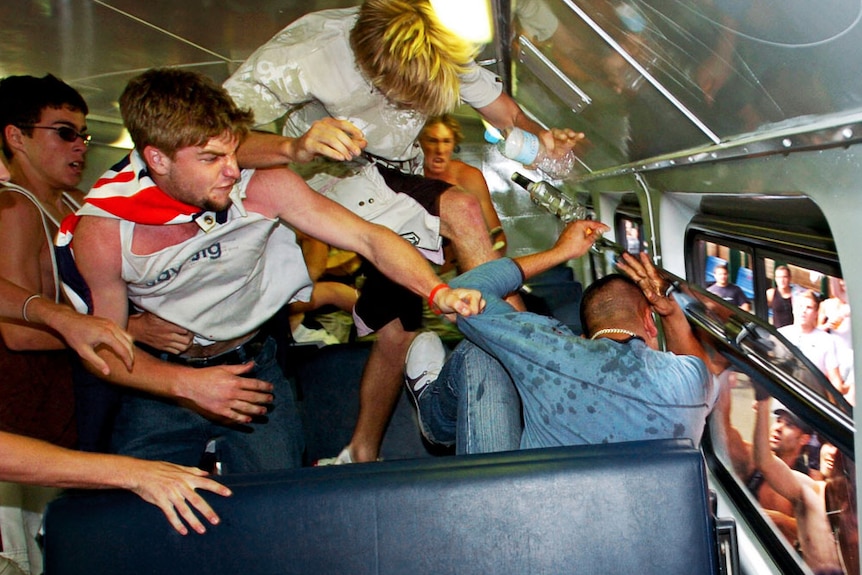 A mob of men, one wearing an flag, rush towards a cowering seated man in static train carriage while a crowd outside watches.