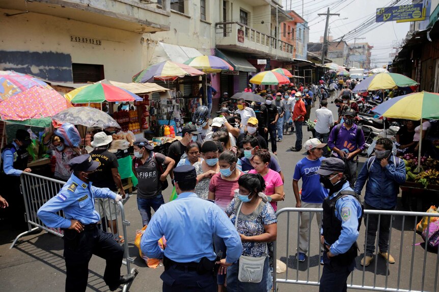A large queue of people wearing face masks wait to be allowed into a market through a pedestrian gate being controlled by police