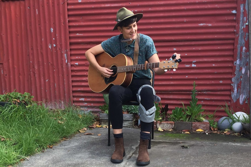 Kerryn Fields sitting in front of a red shed playing her guitar