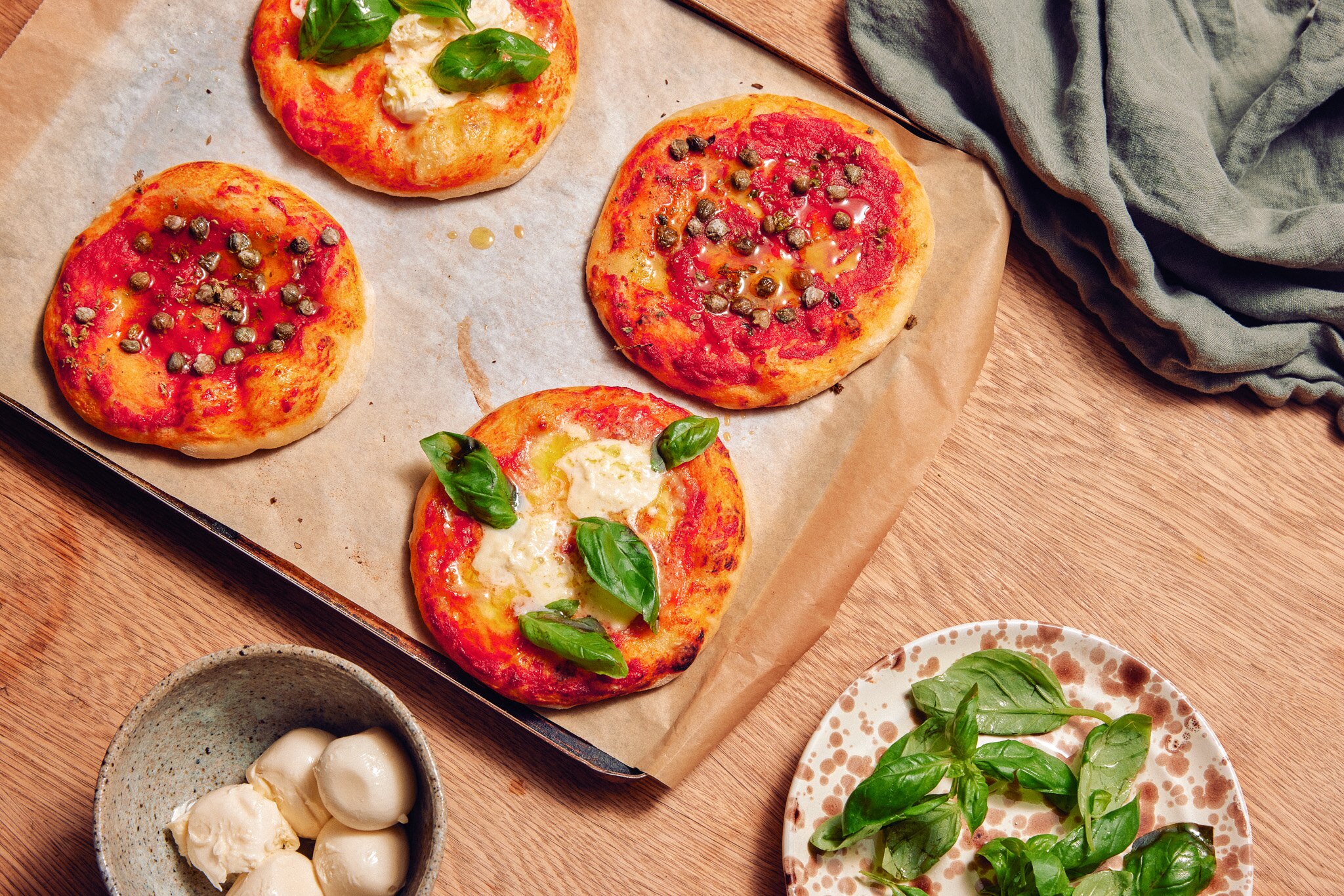 Four mini pizzas on a baking tray with sauce, cheese, basil and capers on them, next to a bowl of basil and cheese.
