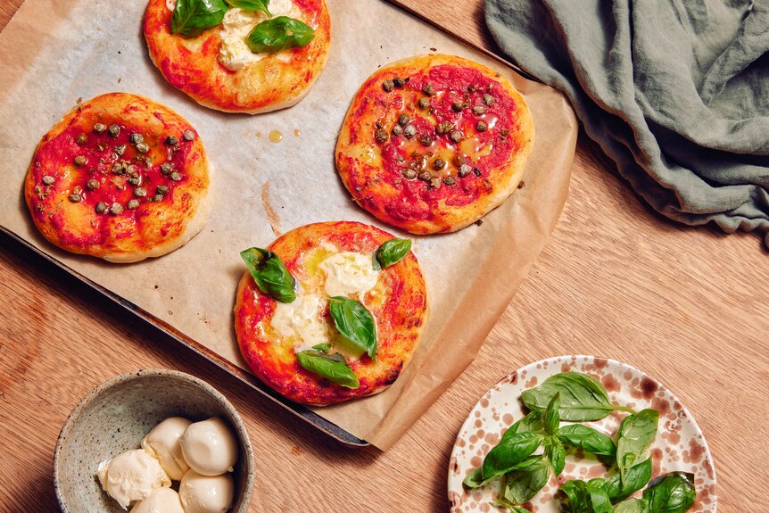 Four mini pizzas on a baking tray with sauce, cheese, basil and capers on them, next to a bowl of basil and cheese.