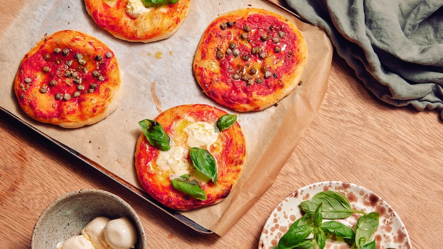 four mini pizzas on a baking tray with sauce, cheese, basil and capers on them, next to a bowl of basil and cheese
