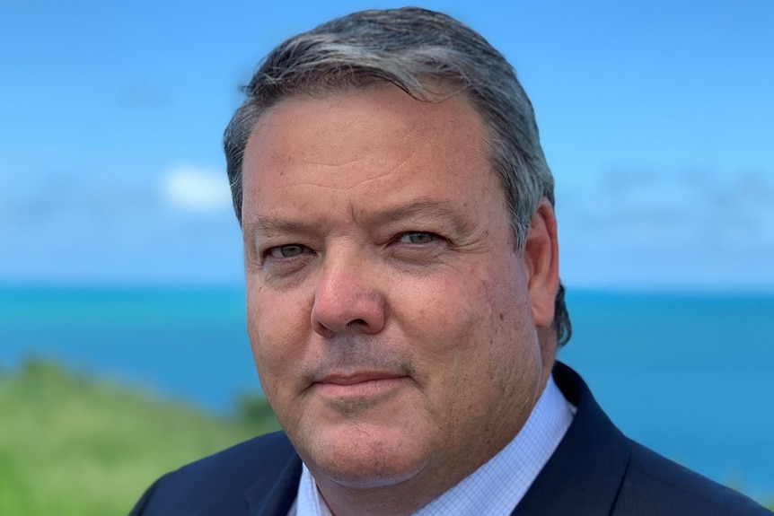 A middle-aged man stands in front of the ocean, looking serious.