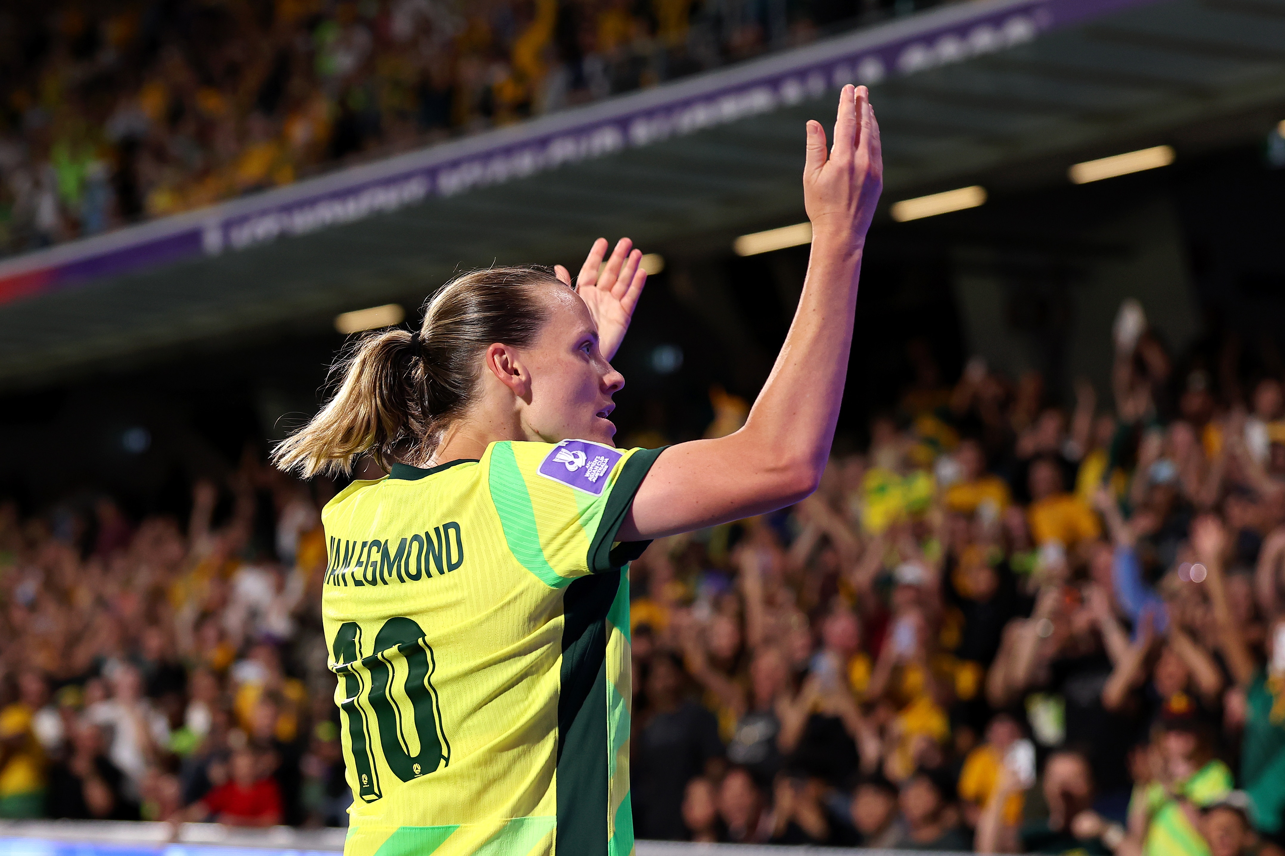 A soccer player in green and gold holds her hands up to the crowd