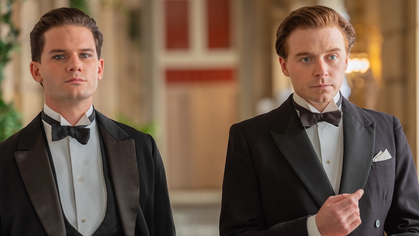 Two young white men dressed in three-piece tuxedos with hair combed back stand in a grand dining room.