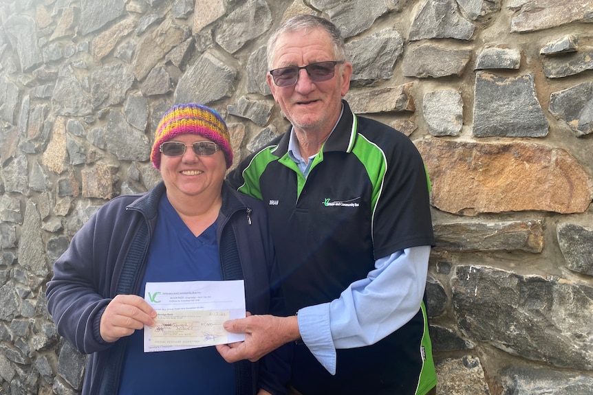 An older woman with a colourful beanie and an older man stand in front of a stone wall, holding a cheque together.