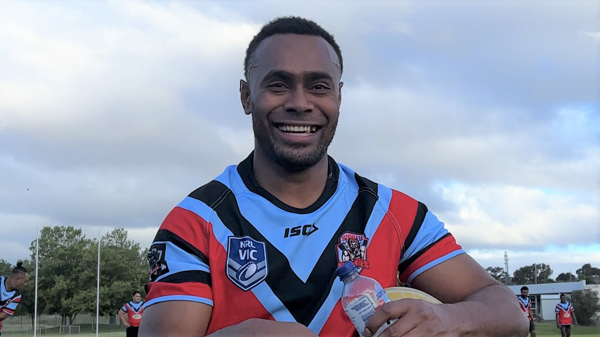 A man wearing a sky blue and red jersey holding a rugby ball and water bottle smiles